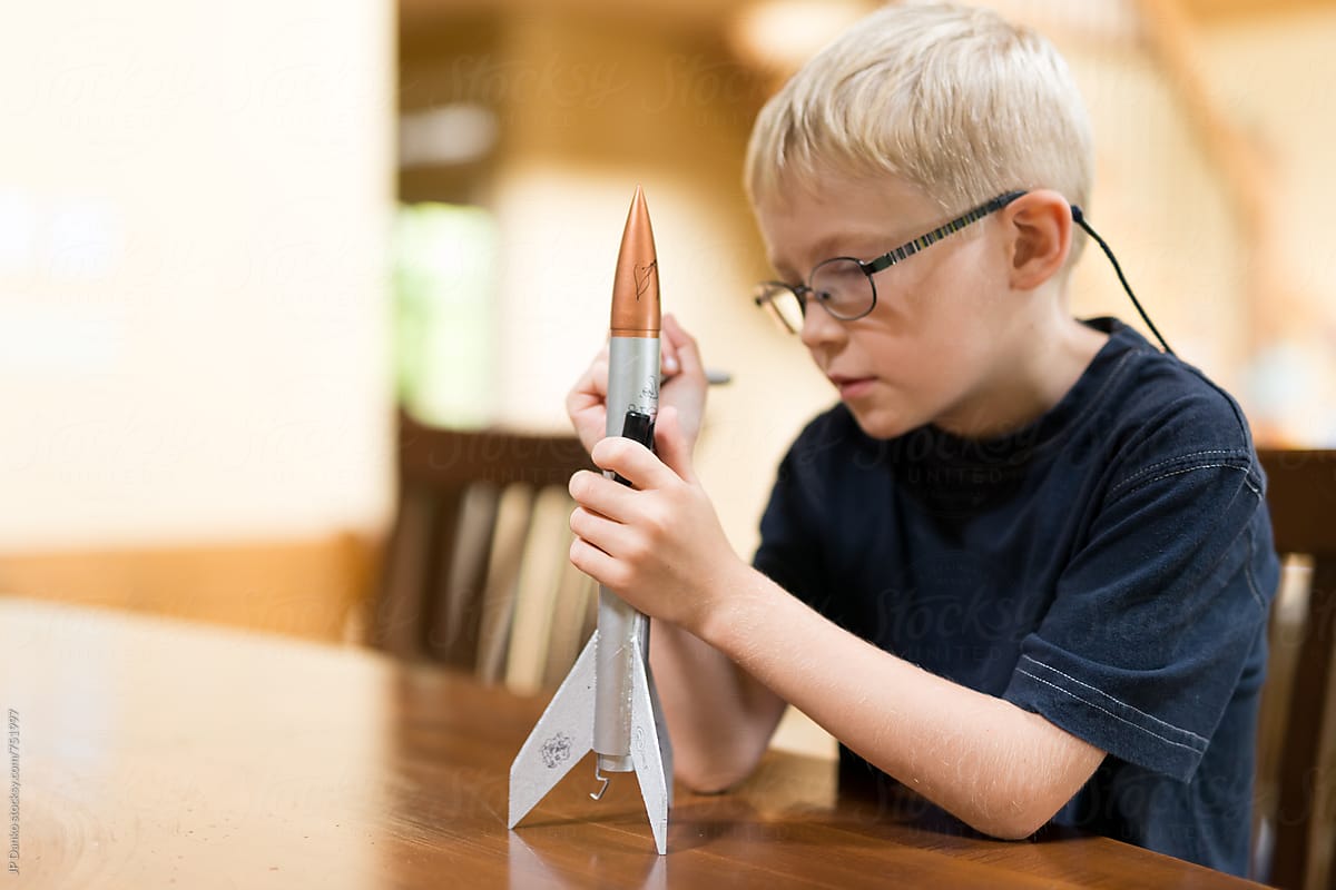 A young boy in glasses sits at a wood table holding a handmade paper rocket, studying it closely, in warm interior light.