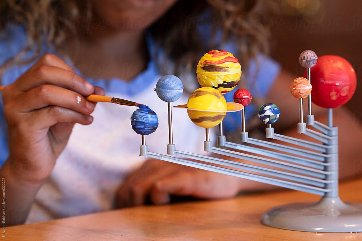 A child painting the planets of a solar system model at a sunlit wooden table.